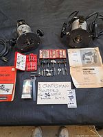 Two Craftsman routers with black tops and metal bases, seen from above, showing detail of handles and router bits included along with manuals and cases.