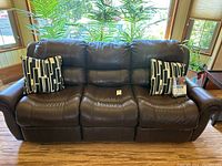 Front view of brown leather reclining sofa with two black and white geometric patterned pillows seated on it, placed in front of windows with indoor plants outside.