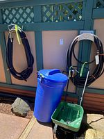 Two black rubber hoses coiled on separate racks mounted on a wood lattice wall next to a blue garbage can and a green Turf Builder fertilizer spreader.