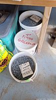 Three white plastic pails filled with different sizes of galvanized nails, with labeled notes describing contents.