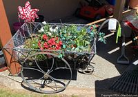 Wide angled photo of the wire wheelbarrow shaped metal planter with artificial flowers and pinwheel decoration, showing the outdoor setting and large spoked wheels.