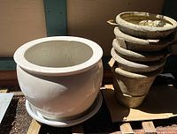 White large fiberglass planter pot with saucer at the bottom, placed outdoors, with a stack of five smaller weathered beige pots to the side.