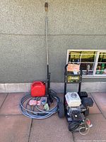 Full view of pressure washer on wheels with hose, wand, box of nozzles and red jerry can on patio against exterior wall