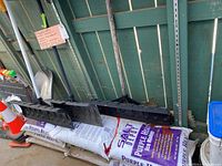 Four snow shovels with black scoops and handles, three appear to be slightly used, one aluminum shovel new, laid on top of stacked ice melt bags.