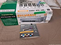 Photo showing Sylvania DVD Recorder box with unit and Roku Streaming Stick+ box in front, labeled 'Never Used' on VCR box.