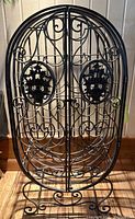 Front view of the black metal wine cage showing decorative floral medallions and scrollwork with double doors closed.