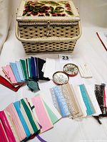 Photo showing woven wicker sewing basket with floral tapestry lid, assortment of colorful seam bindings, lace trims, and metal sewing accessories in tins arranged around basket.