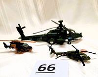 Three die-cast military helicopters arranged on a light-colored surface with a number tag '66'. The large green USAF helicopter, smaller black and tan Matchbox helicopter, and a small army green helicopter are visible.