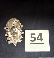 Front view of the Mexican Mayan face pin showing detailed facial features with closed eyes and an elaborate sunray headdress extending upward with scrolls at the sides, placed on a black cloth with a lot number card.
