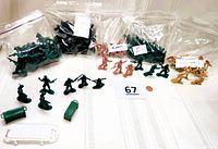 Multiple bags of plastic army men figurines in green, black, tan, and reddish-orange colors displayed on table with handful of loose figures and accessories including green ammo boxes and white plastic crates.