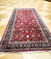 Full view of the large rectangular oriental rug laid out showing maroon field with intricate floral borders and white fringe on the shorter edges.