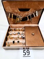 Open men's jewelry box showing tan velvet interior, ten tie clasps secured on a band across upper lid, assorted cuff links and tie tacks in slots below, and closed button-closure compartment on right side of the tray.