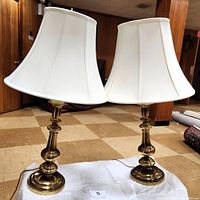 Pair of brass candlewick style lamps side by side showing full bodies and white bell shades on tiled floor indoors.
