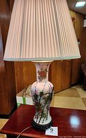 Full view of vintage Asian-style porcelain lamp on wooden base with white pleated shade; placed on wooden table against paneled wall.
