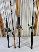 Three fishing poles standing vertically with attached reels: blue foam grip, cork handle, and black foam grip types.