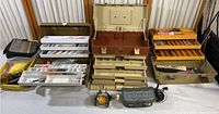 Photo showing three fishing tackle boxes open with visible fishing lures, tackle trays, and a fishing reel.
