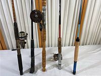 Photo showing five fishing poles standing upright on table, various handles and reels visible.