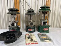 Three vintage Coleman green fuel lanterns with glass globes and metal tops. Includes black metal funnel and unopened replacement parts packaged in blister packs.