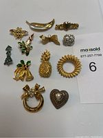 Eleven vintage brooches arranged on a white background including fruit, bows, hearts, and floral designs, some accented with rhinestones and pearls.