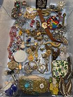 Top down view of a clear container filled with an assortment of vintage costume jewelry and craft making items, including a brown leather wristwatch, gold-tone brooches, pearl necklaces, and various pins and pins with Disney themes.