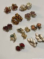 Photo showing 14 pairs of vintage clip-on earrings arranged on a white surface with various colors and designs including faux pearls, rhinestones, and enamel.