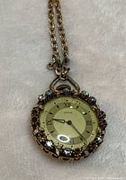 Front view of vintage ladies pocket watch showing rhinestone studded bezel, gold-tone face with roman numerals, and attached chain.