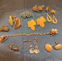 View of vintage jewelry collection laid out on dark fabric background including earrings, bracelets, and brooches with varying designs in gold-tone metal and colorful accents.