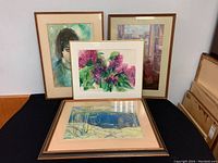 Four framed artworks leaning against wall on floor showing the two main artworks in front with lake scene and floral print.