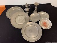Photo of eight pewter pieces including candleholder, pitcher and six plates/dishes on black fabric background.