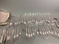 Top-down view of multiple flatware sets in clear plastic sleeves, showing spoons, forks, and knives with polished silver finish.
