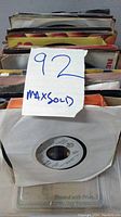 Top view of multiple 7-inch 45 RPM vinyl records in sleeves stacked vertically, showing the edges of many record sleeves with a handwritten sign '92 MaxSold'.