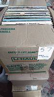Box filled with vinyl record albums seen from above with some spines and cover edges visible. The box is sealed taped and marked U-Haul, labeled with lot number 57 from MaxSold.