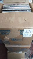 Stack of approximately 100 vinyl records in a cardboard box labeled with MaxSold sticker '29'. The box is worn.