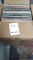 Box with approximately 25-30 vinyl LP records stacked, showing condition and quantity.