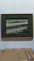 Front view of the framed Japanese woodblock print showing Mount Fuji, a boat, and figures below, surrounded by dark gray fabric mat and gold frame.