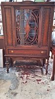 Front view of antique wooden tall cabinet with glass door featuring decorative wood lattice, drawer below, turned legs, and lower shelf.