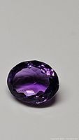 Oval cut natural amethyst gemstone with deep purple color and bright reflections, lying on neutral background.