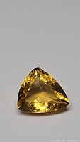 Close up photo of a natural golden citrine gemstone with a trillion cut, showing its facets and clarity on a gray background.