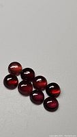 Photo showing eight round cabochon garnet stones laid out on a gray surface, displaying deep red colors and reflective luster.