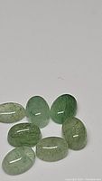 Seven oval cabochon cut green kyanite gemstones spread on a white background, showing their translucent quality and internal textures.