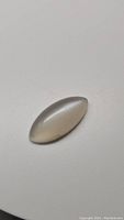 Single marquis cabochon cut white silver moonstone showing cat's eye flash on plain white background.
