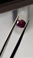 Photo showing the first oval cut natural sapphire gemstone held by tweezers, showcasing deep reddish pinkish purple color and some internal inclusions.
