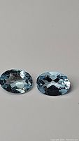 Pair of oval-cut sky blue topaz gemstones lying flat on a neutral background, showing clarity and color.