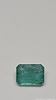 Rectangular emerald-cut natural emerald gemstone photographed on a grey background, showing its green color and typical internal inclusions.