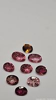 Top-down clear image showing 10 oval cut pink tourmaline gemstones arranged with one darker stone near the top.