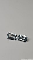 Pair of rectangular emerald-cut light blue topaz gemstones shown on flat white surface with clear faceting and reflections.