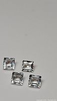 Four clear square cut white topaz gemstones on a light surface showing reflections and polished facets.