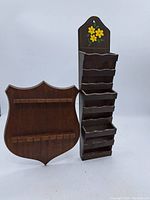 Photo showing the dark wooden letter sorter with six scalloped slots and hand-painted flowers, alongside the shield-shaped wooden spoon holder with two rows of hooks.