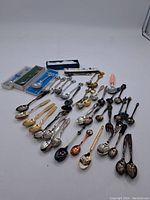 Photo displaying the full assortment of 34 souvenir spoons in multiple finishes including silver-tone, gold-tone, with some enamel decoration. Various sizes and designs visible, some in plastic cases.