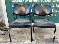 Pair of black metal chairs showing front angle, seats and backs visible, placed on concrete with sign in background